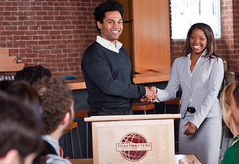 Man and woman shaking hands at a lectern.  The lectern has a Toastmaster logo on the front